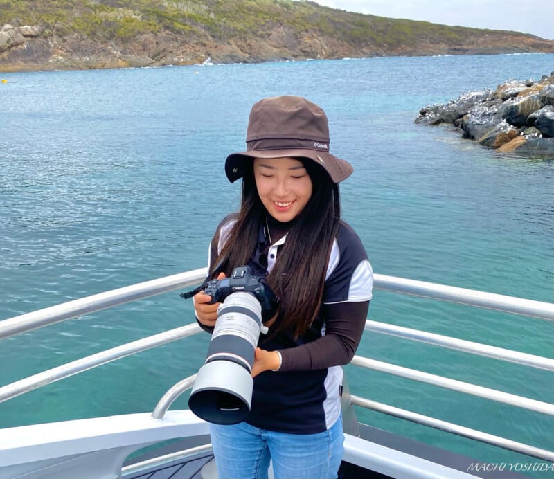A smiling woman wearing a bucket hat holds a camera with a large lens on a boat, with a coastline and calm sea in the background.