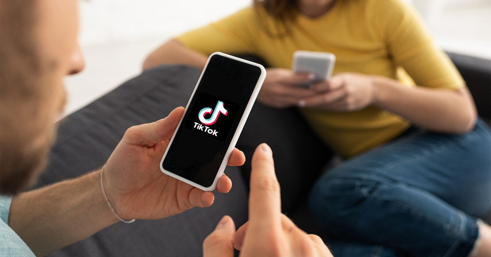 A man and a woman sitting on a sofa, each using their smartphones. the man's phone, held in his hand, displays the tiktok logo on its screen. focus on the man's phone screen.