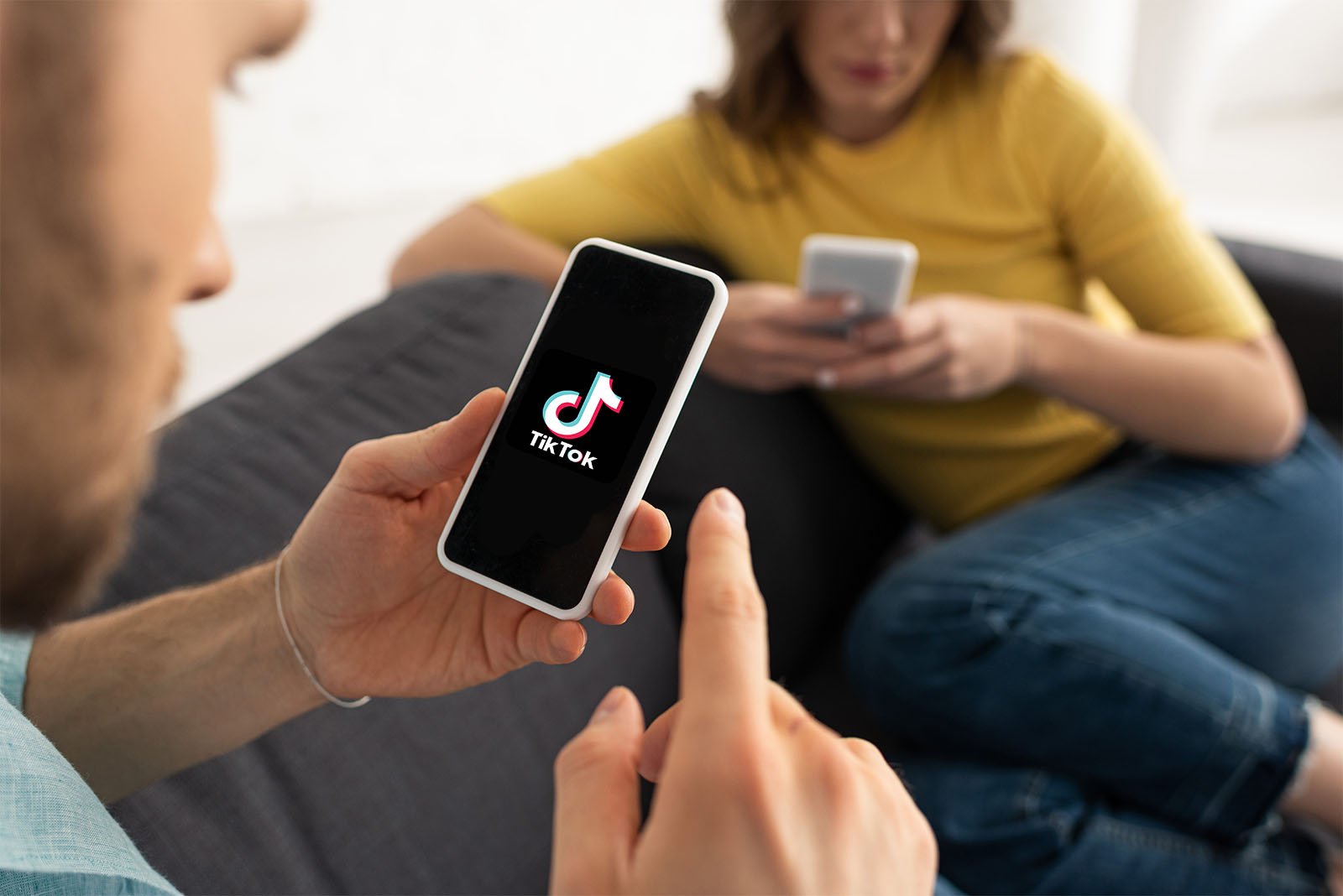 A man and a woman sitting on a sofa, each using their smartphones. the man's phone, held in his hand, displays the tiktok logo on its screen. focus on the man's phone screen.