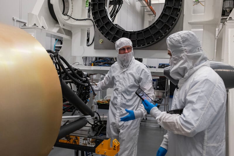 Two scientists in cleanroom suits inspect equipment and a large spherical object in a high-tech laboratory setting.
