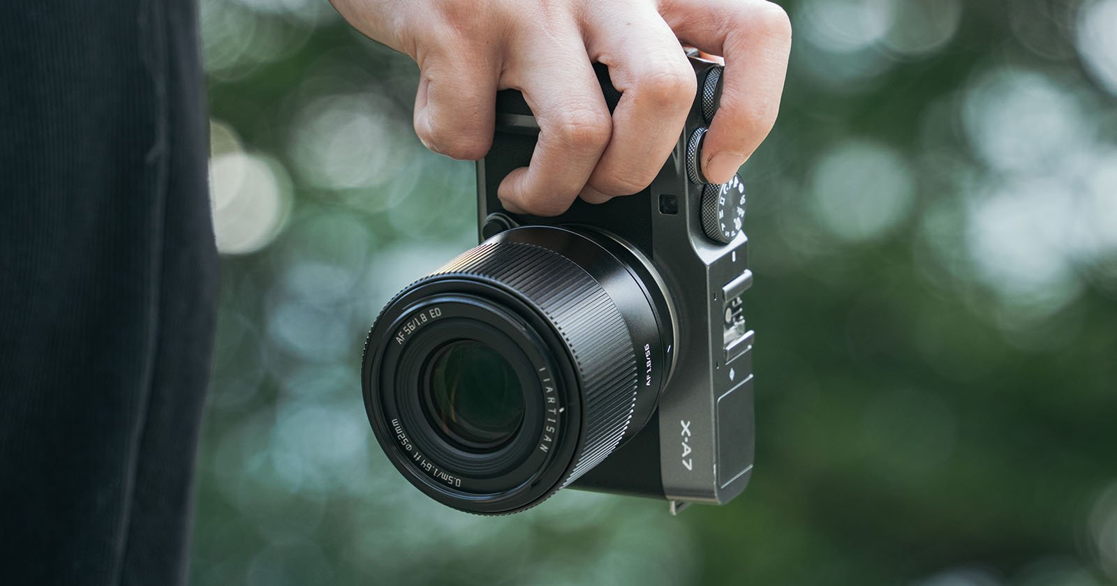 A close-up of a person's hand holding a modern camera by its strap, with a blurred green background. the camera is oriented with the lens facing forward.