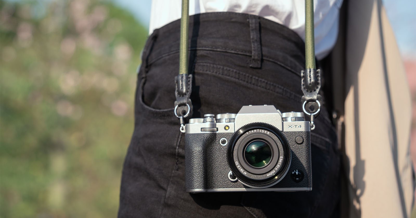 A close-up view of a vintage-style silver mirrorless camera with a black strap, hanging from a person's belt against a blurred natural background.