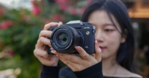 A woman with dark hair holding a digital camera up to her face, focusing it. the setting is outdoors with soft-focus greenery in the background.