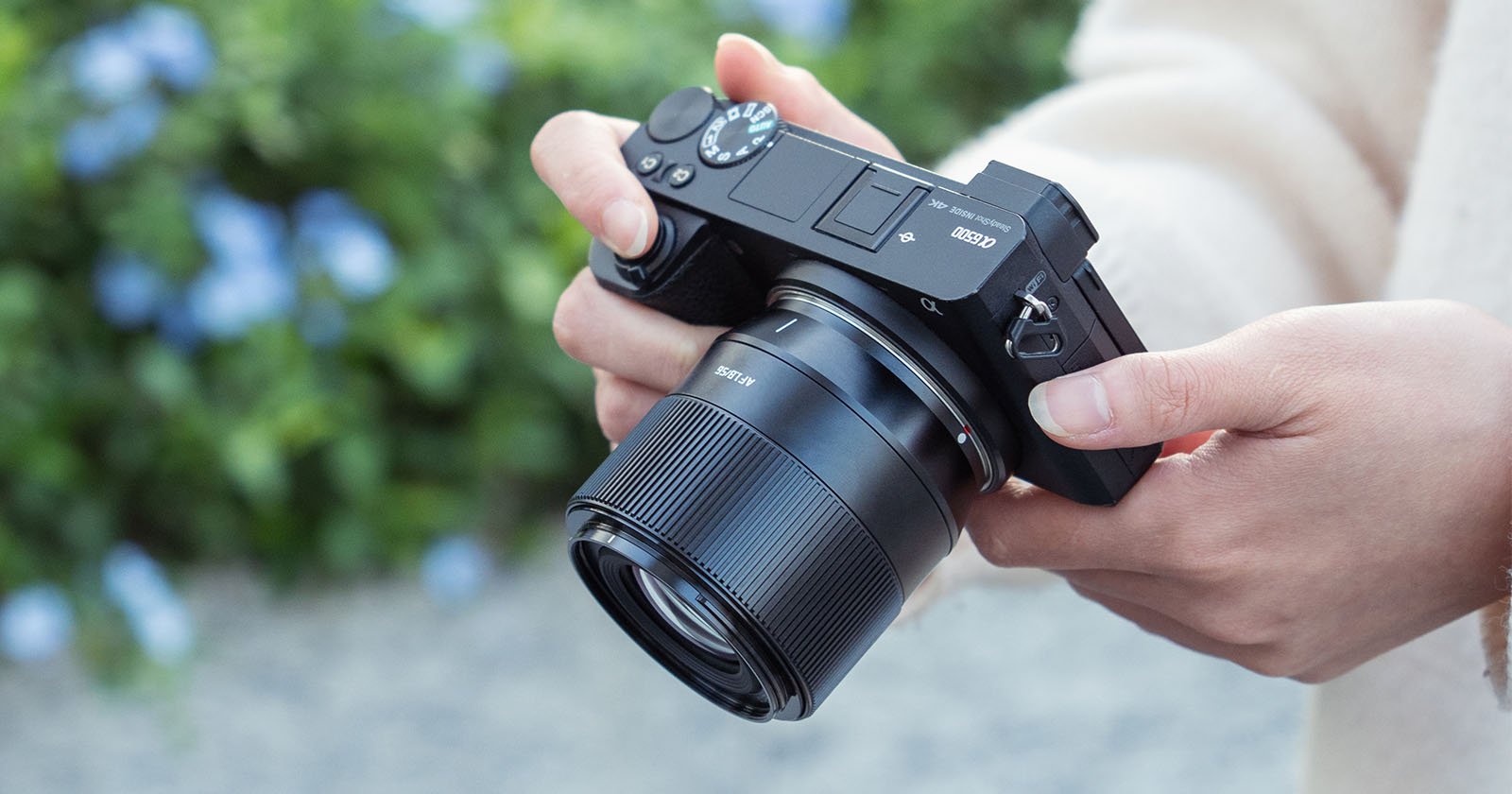 A person holding a black dslr camera, adjusting settings with their right hand, against a blurred green foliage background.