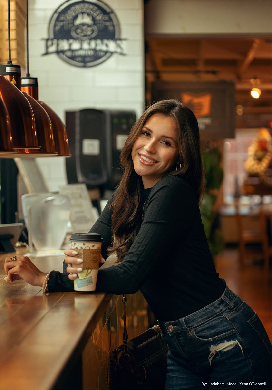 A woman with long brown hair, smiling and holding a coffee cup, leans on a cafe counter under warm lighting, with a decorative emblem in the background.