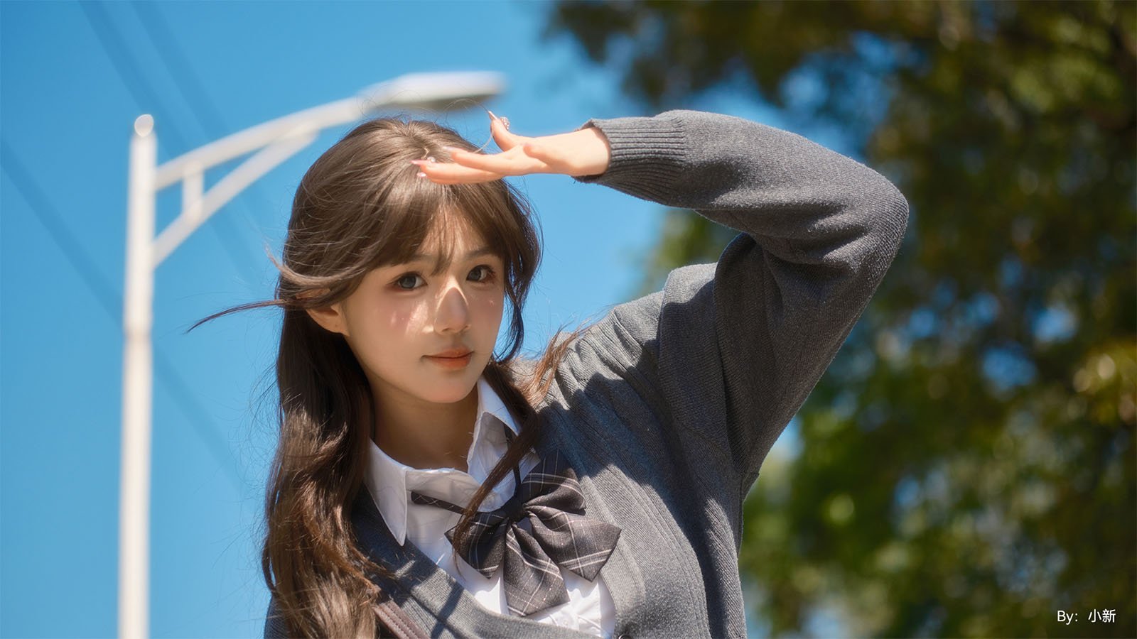 A young woman in a school uniform salutes the camera with her hand above her eyes, against a backdrop of clear blue skies and blurred greenery.
