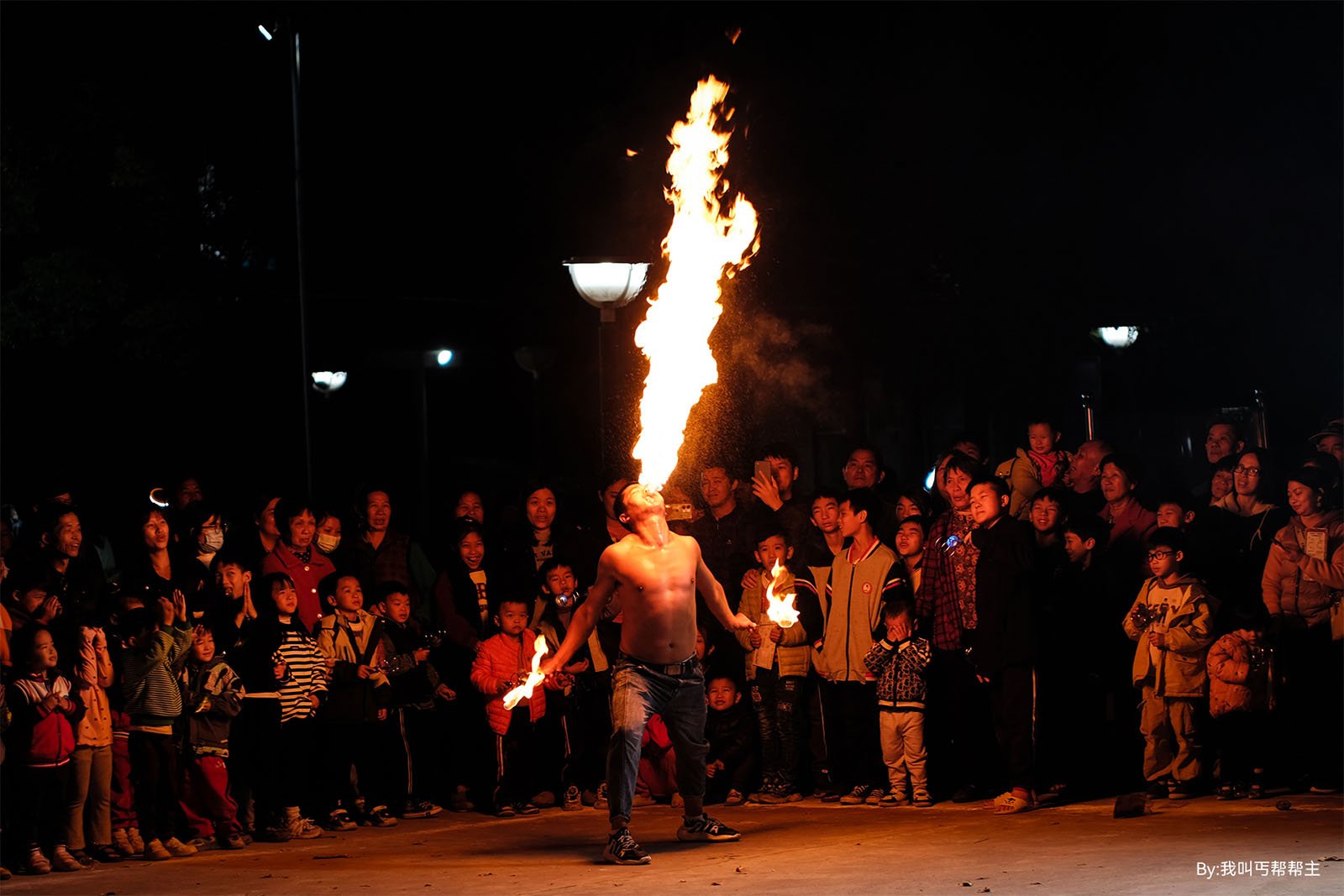 A shirtless fire performer breathes a large flame into the night sky, illuminated by a street lamp and surrounded by an amazed crowd.