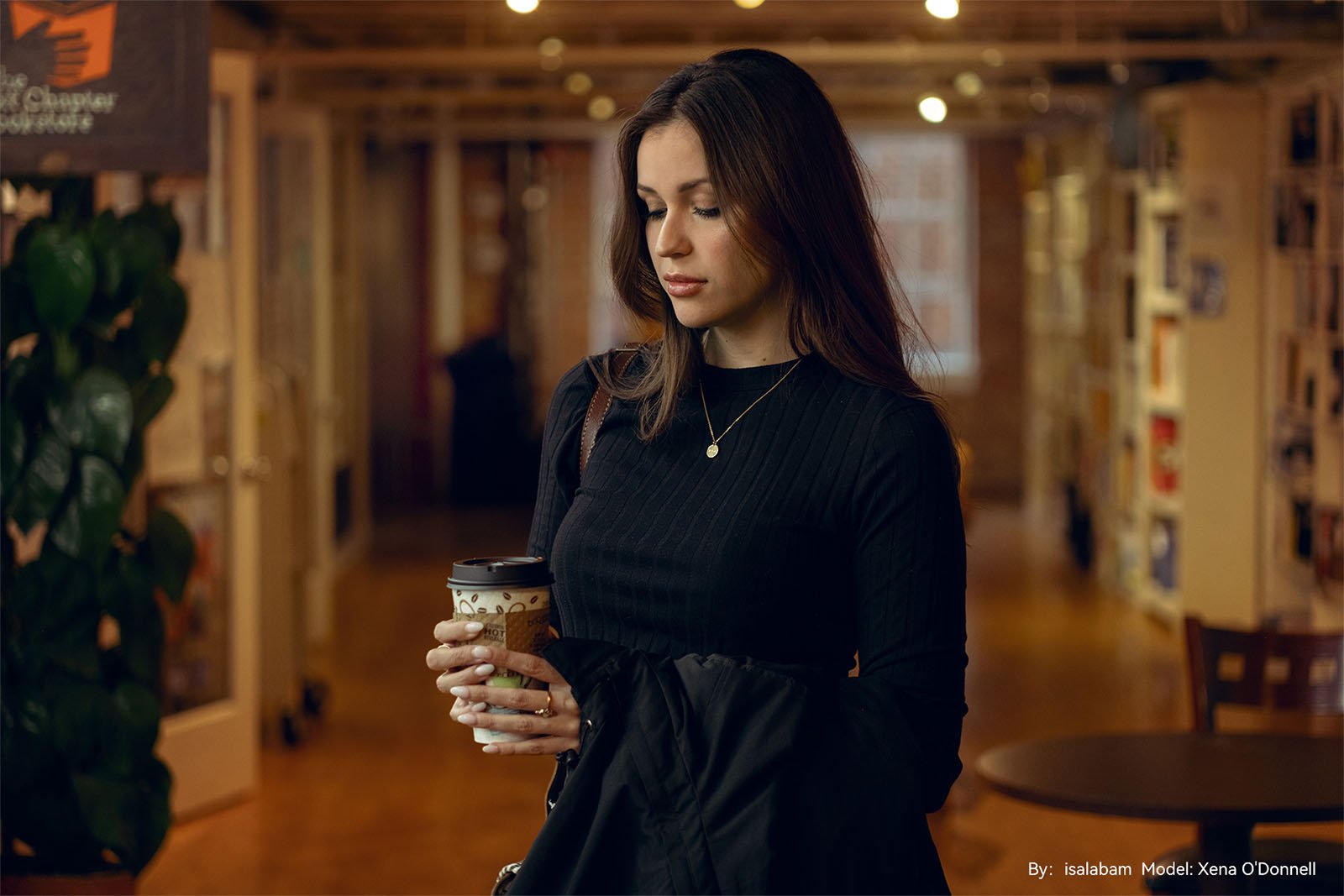 A woman in a black sweater holds a coffee cup and a jacket inside a warmly lit cafe with blurred background showcasing indoor plants and hanging lights.