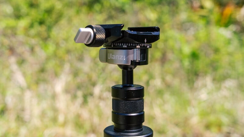 A close-up of a ulanzi camera tripod head, featuring a ball head and adjustment knobs, set against a blurred green natural background.