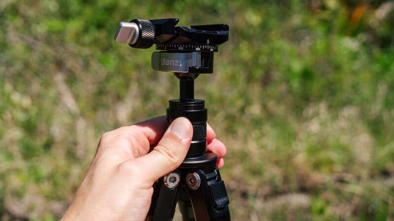 A hand holds the Ulanzi trekking tripod in front of a blurred grassy background. 