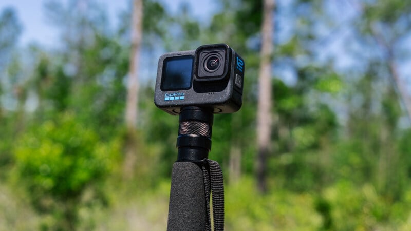 A gopro camera mounted on an Ulanzi selfie stick, centrally framed against a blurred forest backdrop under clear blue skies.