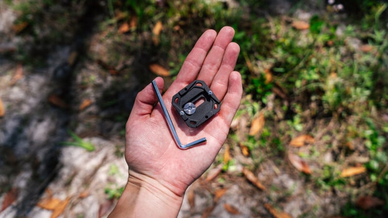 A person's hand holding a small, black camera tripod mount and an allen wrench outdoors, with a natural, leaf-covered ground background.