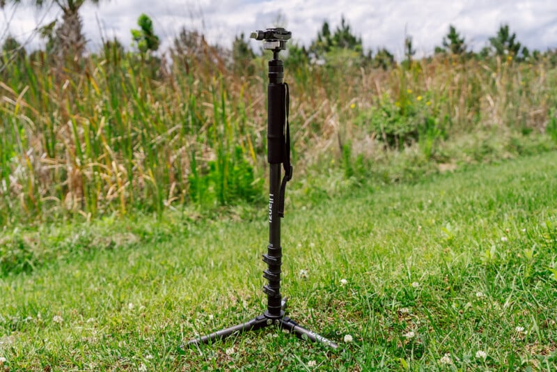 A tripod stands on a grassy field with a blurred background of green vegetation and a partly cloudy sky.