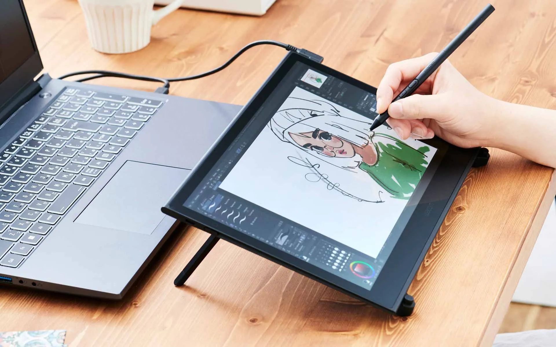 A person's hand using a stylus on a digital drawing tablet that's connected to a laptop, working on a cartoon illustration of a girl with blonde hair.