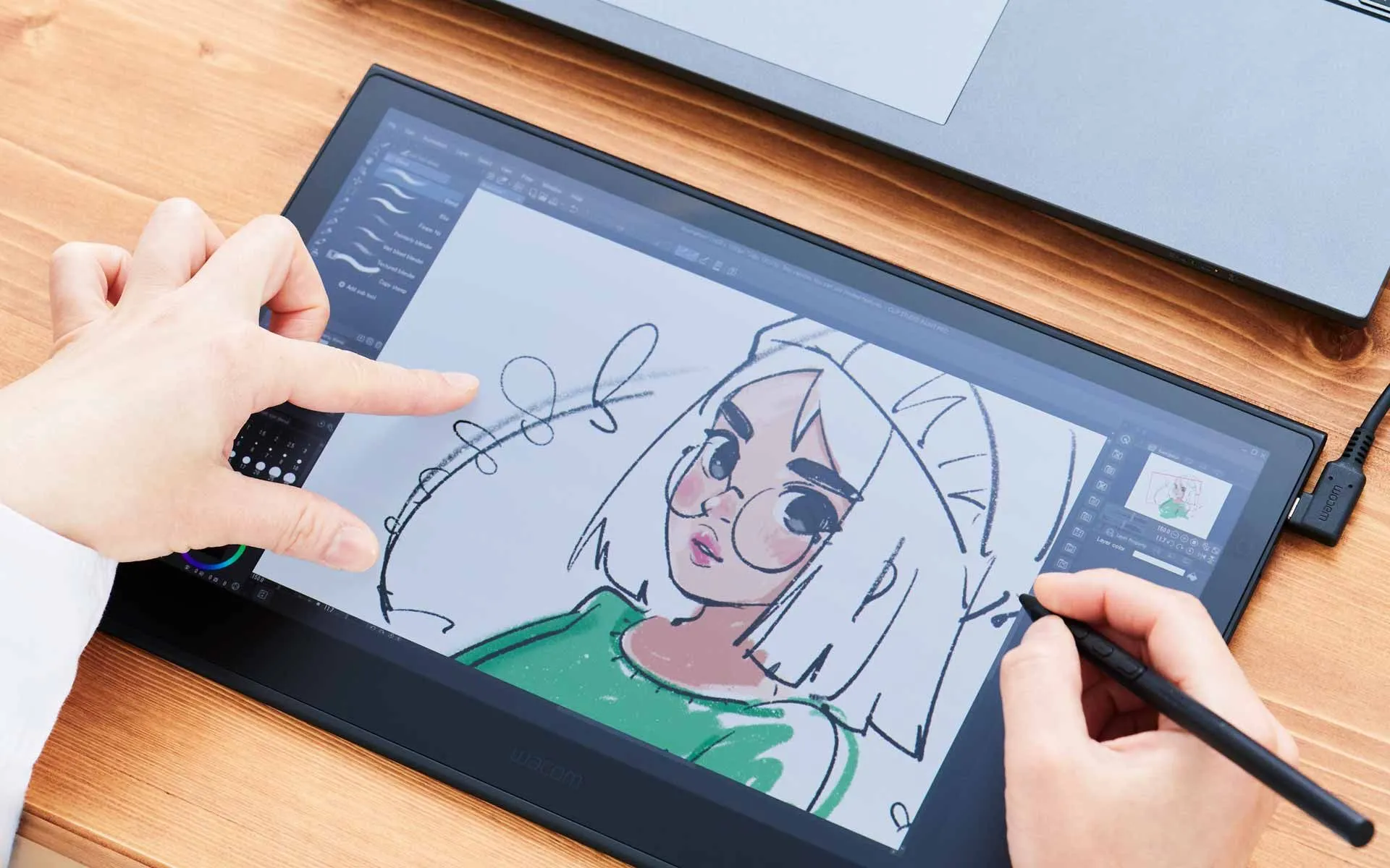 A person uses a stylus on a digital drawing tablet to create a colorful illustration of a female character, with a laptop and papers visible on a wooden desk.