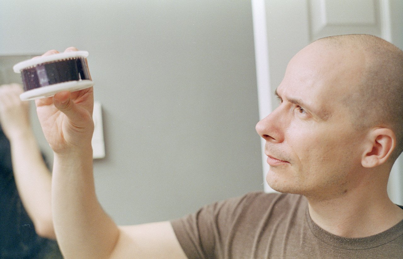A bald man intently examines a strip of film negatives he is holding up to the light in a room with a light blue wall.