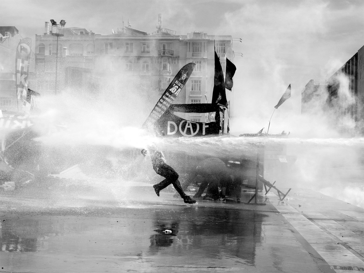 A black-and-white photo of a person running through a water cannon spray during a protest, with banners waving and urban buildings in the background.