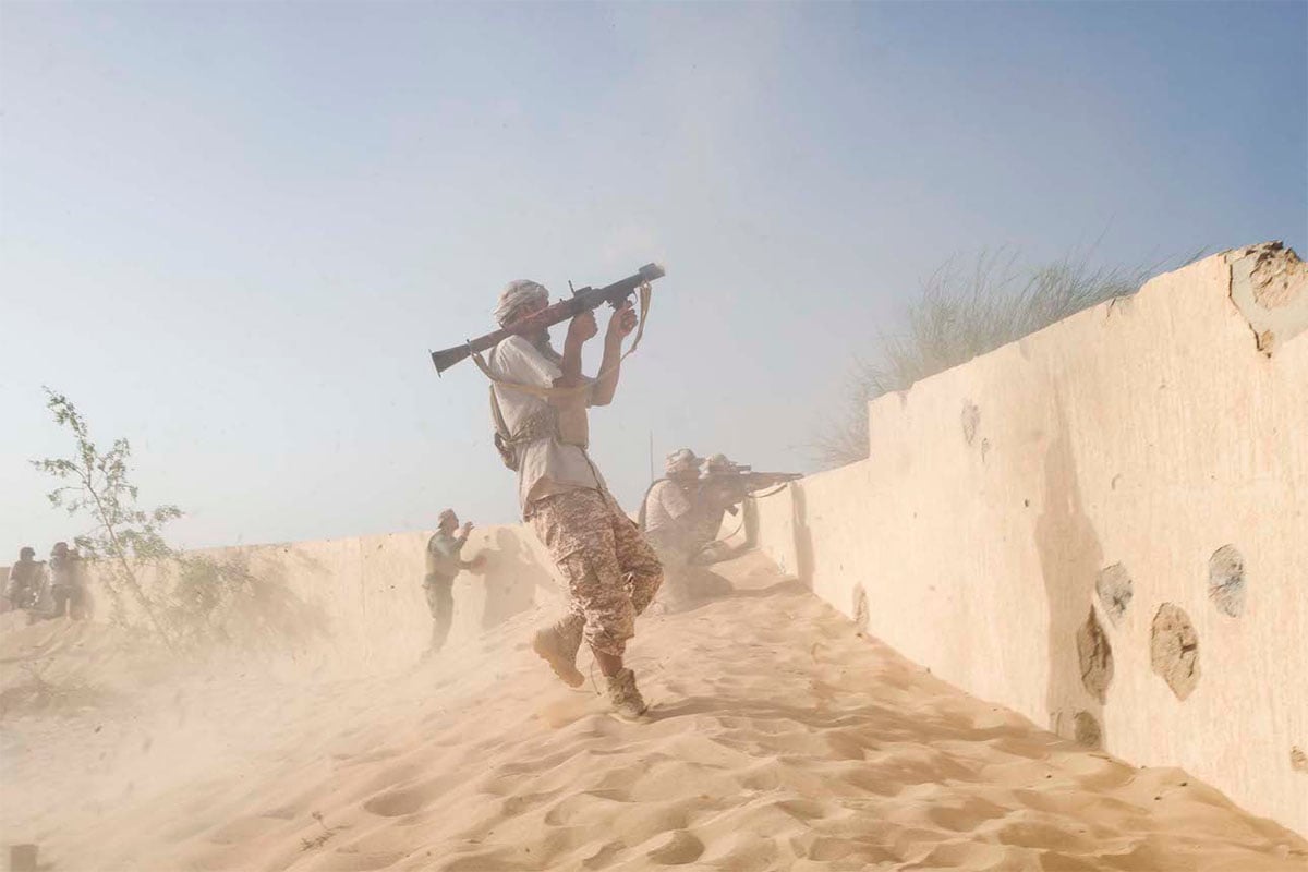  Soldiers in camouflage uniforms practice an assault, firing weapons amid a dusty, sandy environment with a clear sky overhead.