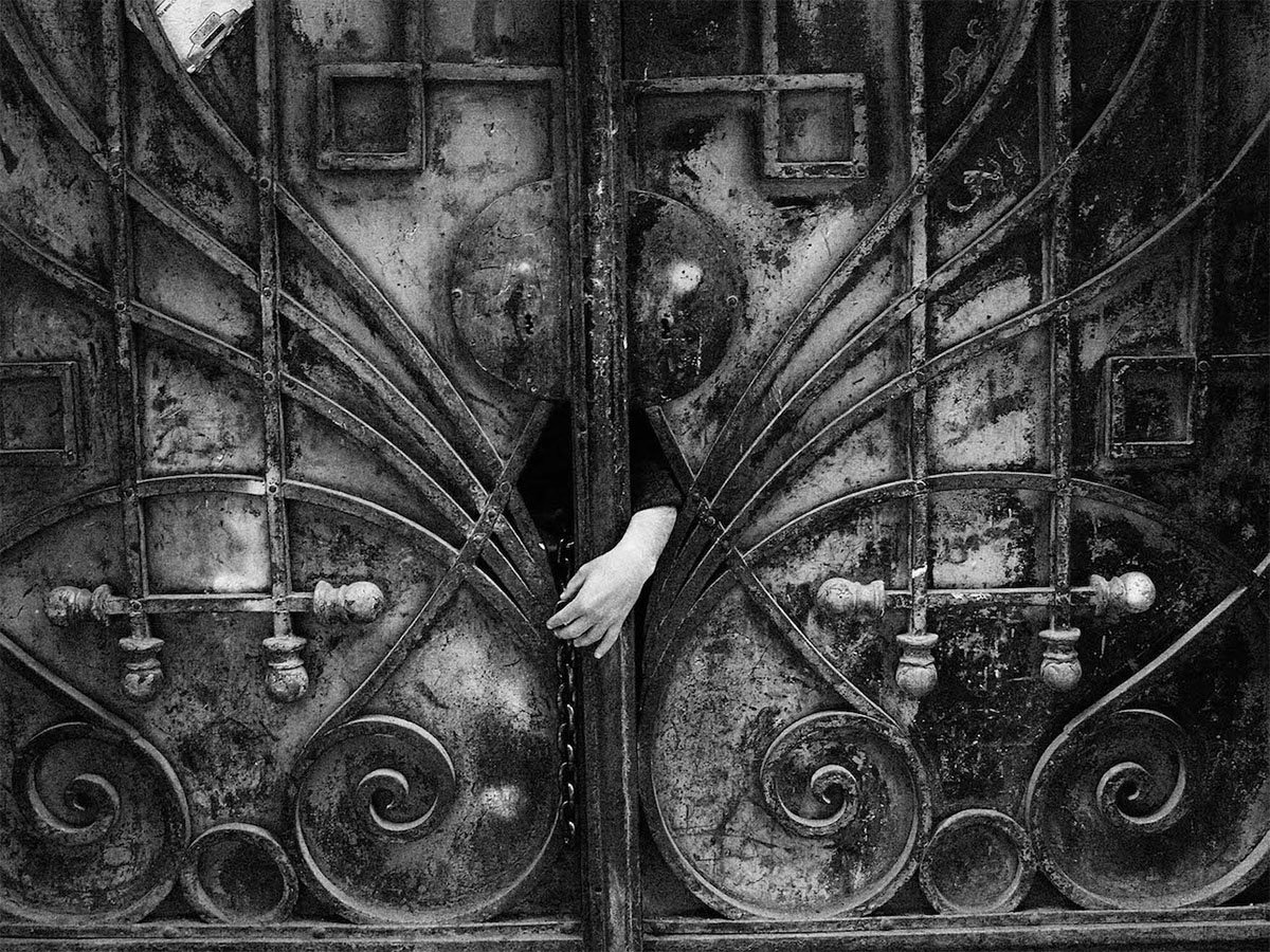  A black and white image of a vintage, ornate metal door slightly ajar with a human hand visible between the opening. the door features intricate patterns and textures.