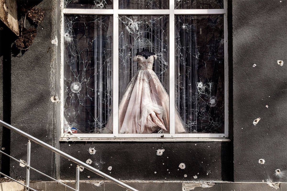A wedding dress displayed in a bullet-riddled window of a damaged building, symbolizing stark contrast between beauty and destruction.