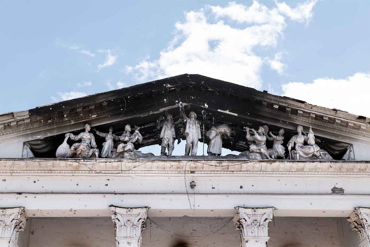  Weathered neoclassical pediment with sculptural figures on a building, showing signs of damage and decay against a clear sky.