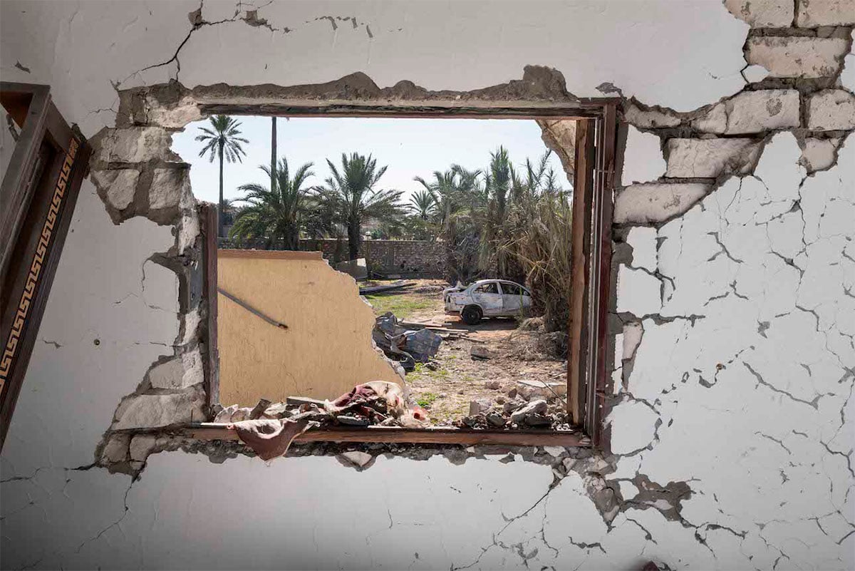  View through a damaged wall opening showing a derelict urban scene with a wrecked car, scattered debris, and palm trees in the background.