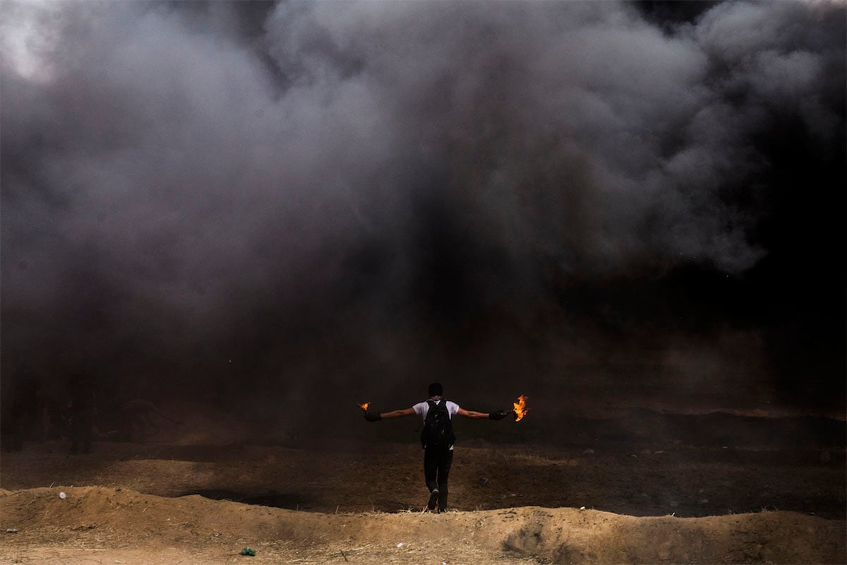 A person with outstretched arms holding flaming objects stands in a smoky, desolate landscape, emphasizing a dramatic or protest-like scene.