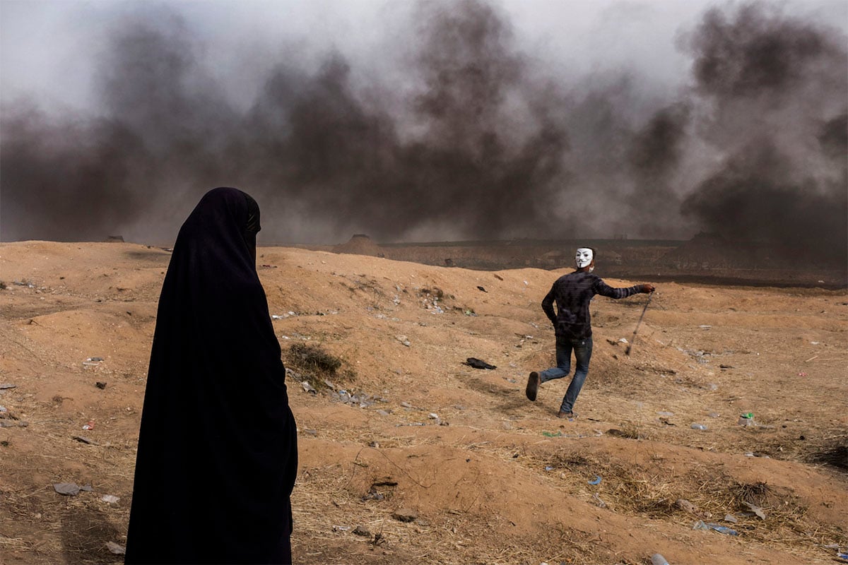 A woman in a black hijab stands to the left, watching a man with a painted face and dark clothing run through a smoky, barren landscape.