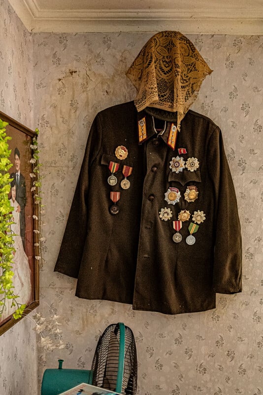 A military jacket decorated with various medals and ribbons is hanging on a wall. A lace shroud covers the top part. There's a framed photo of a couple in wedding attire on the left side and the top part of a fan visible at the bottom. The wall has a floral pattern.