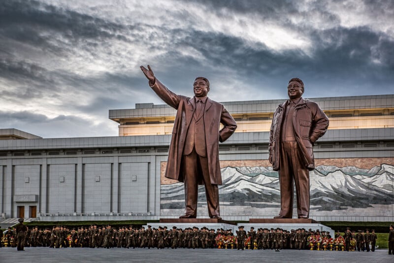 A large bronze statue of two men stands in front of a building with a mural of mountains. One man is waving while the other has his hands at his sides. A crowd of people is gathered at the base of the statues, with many of them in military uniforms. The sky is overcast.