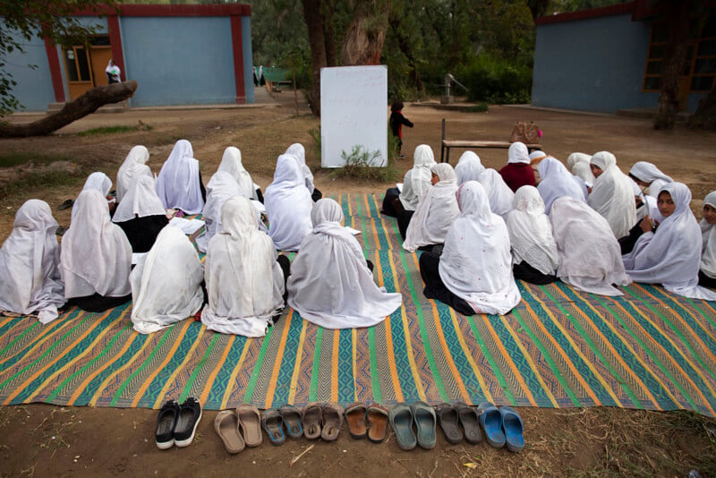 A group of girls, all wearing white headscarves, sit on a colorful mat outdoors, facing a whiteboard with writing on it. A row of sandals is neatly lined up in the foreground. Two blue buildings and trees are visible in the background.