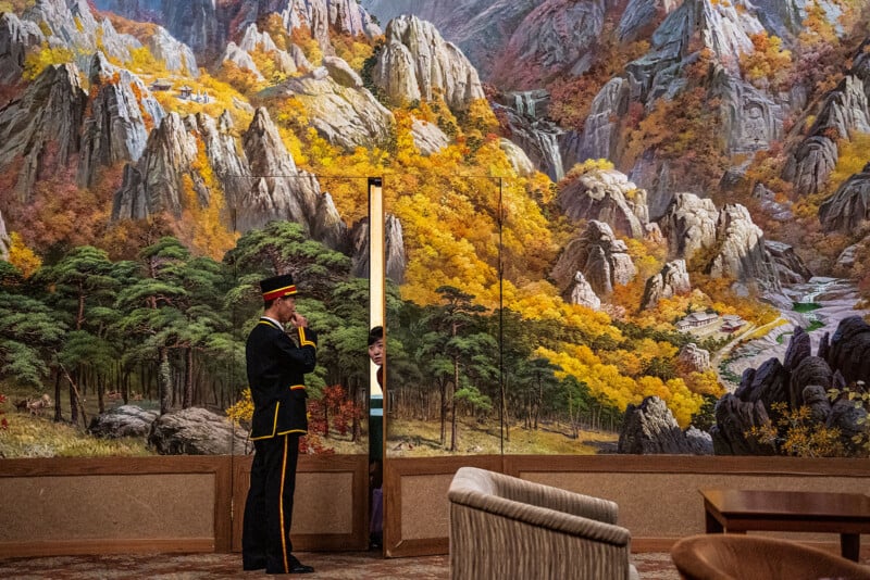 A person in a formal uniform stands by a door, partially opening it to reveal another person peeking through. The background is a vibrant mural depicting a mountainous landscape with trees in autumn colors. Various furniture pieces are in the foreground.