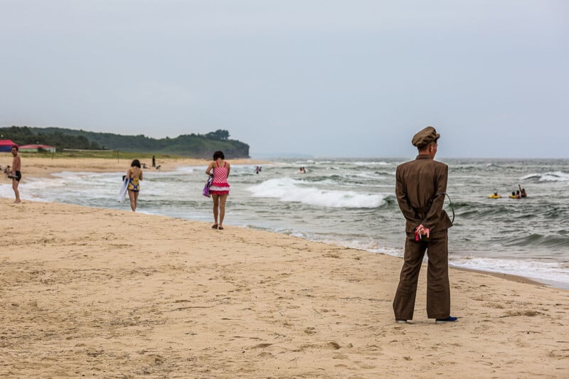 A person in uniform stands on a sandy beach with hands behind their back, looking towards the ocean. Several beachgoers, some in swimwear, are seen walking along the shore and playing in the water. The sky is overcast, and a forested area is visible in the background.