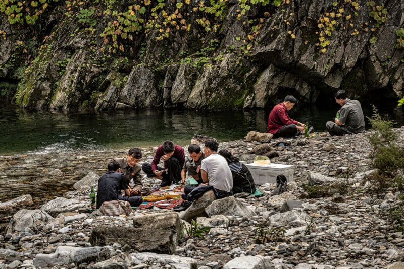 A group of people are sitting on rocky ground by a river, enjoying a picnic. They are surrounded by lush greenery and large rock formations. Food items and picnic supplies are spread out on blankets in front of them.