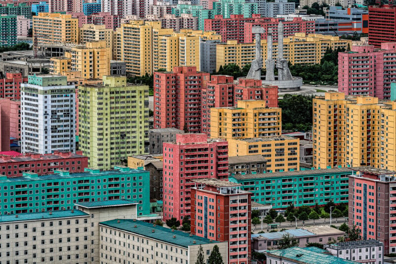 A densely packed cityscape featuring numerous colorful high-rise apartment buildings in shades of yellow, pink, green, and blue. In the background, a unique monument with three large, sculptural towers is visible amidst the buildings.