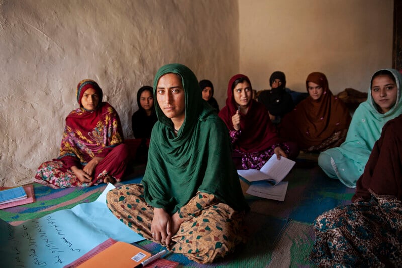 A group of women sit on the floor in a modestly furnished room. They are dressed in colorful traditional clothing and scarves. Several hold notebooks and papers. The woman in the foreground, in a green headscarf, looks directly at the camera with a serious expression.