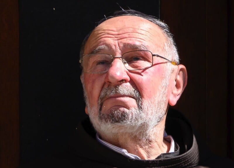 An elderly man with a white beard and glasses looks upward with a contemplative expression. He is wearing a dark robe and is set against a dark background. The sunlight casts a warm glow on his face.
