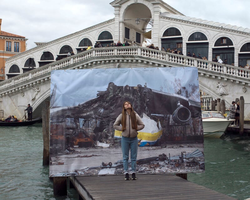 A person stands on a dock by a canal, with the Rialto Bridge in the background. They are looking up, and behind them is a large photographic display showing the wreckage of a building. Many people are on the bridge, and several boats are visible on the water.