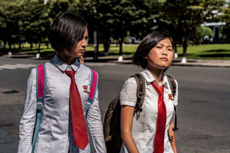 Two young students wearing white shirts, red ties, and backpacks walk on a sunny street. One has a bright blue backpack, and the other has a beige backpack. Both have badges pinned to their shirts. Trees and a grass area can be seen in the background.