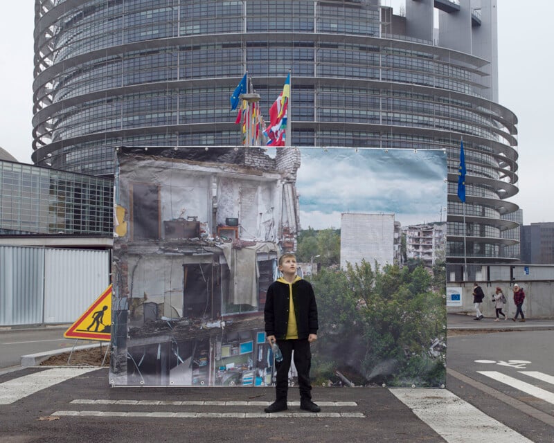 A boy stands on a street, looking up at a large poster depicting a war-torn building. The modern, curved architecture of a different building is visible behind the poster. Flags and pedestrians are also seen in the background.