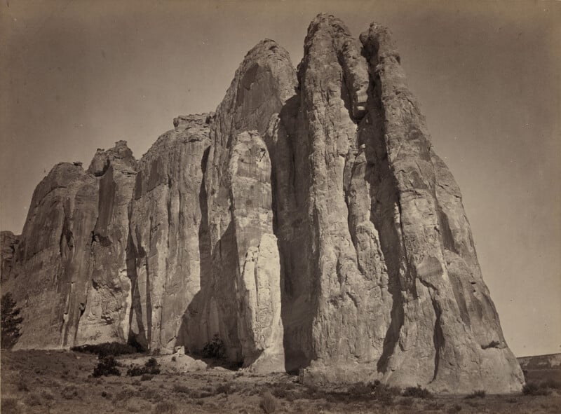 A black and white photograph of a towering rock formation with multiple vertical columns and rugged surfaces. The ground is covered with sparse vegetation, and the sky is clear with no clouds visible. The landscape appears arid and expansive.