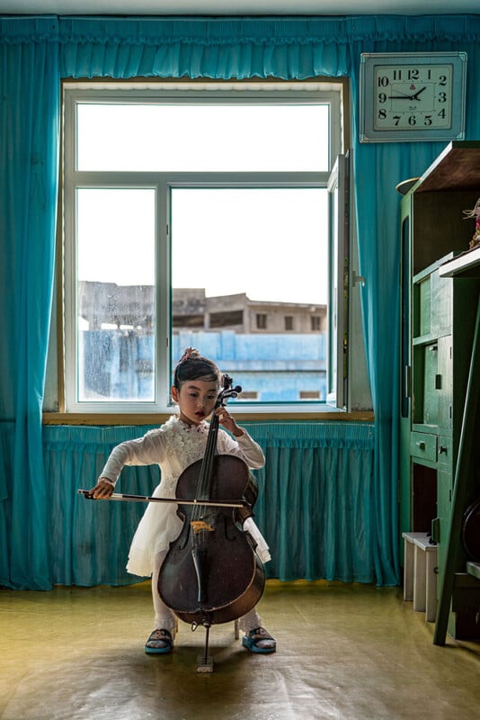 A young girl in a white dress plays a cello in a room with blue curtains and a wall clock showing 3:55. She is seated near an open window that shows part of a building outside. The room appears orderly with a green piece of furniture on one side.