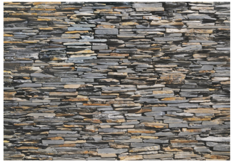 A close-up image of a wall constructed from unevenly stacked, rectangular, flat stones in various shades of grey and brown. The stones are tightly fitted together, creating a dense, textured pattern.