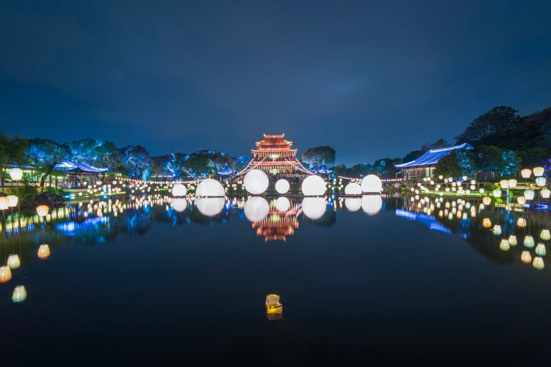 A serene nighttime scene of a traditional chinese garden with a pagoda, illuminated with bright lights and lanterns reflecting on a calm lake.