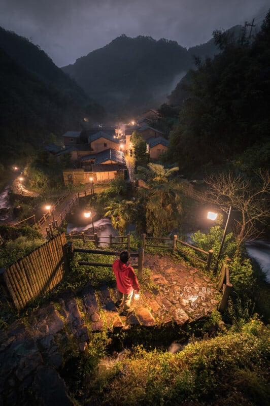 A person in a red cloak stands overlooking a misty, lamplit village nestled in a valley at twilight, with a river flowing alongside. the scene exudes a mystical, serene atmosphere.