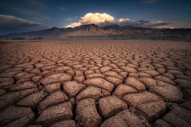 A cracked dry riverbed in the foreground with distant mountains and cloudy skies during sunset, capturing a dramatic and arid landscape.