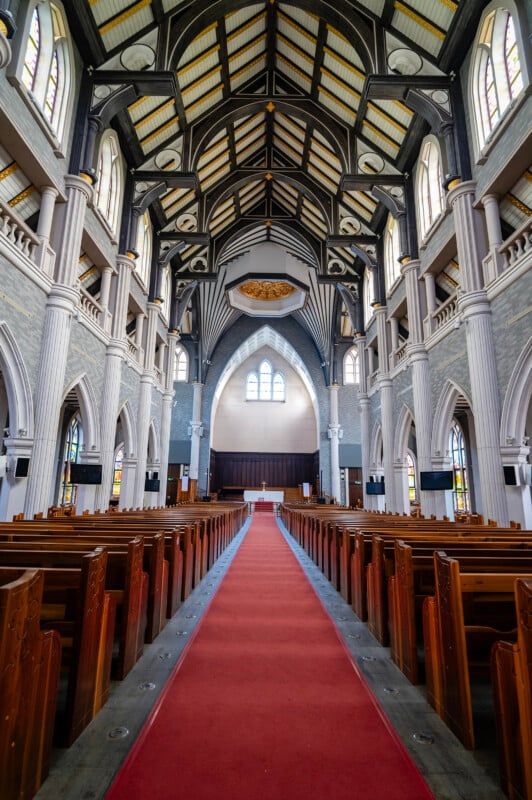 Interior of a church with a vaulted ceiling, wooden pews aligned on a red carpet, and tall arching white pillars leading to a large window at the front.