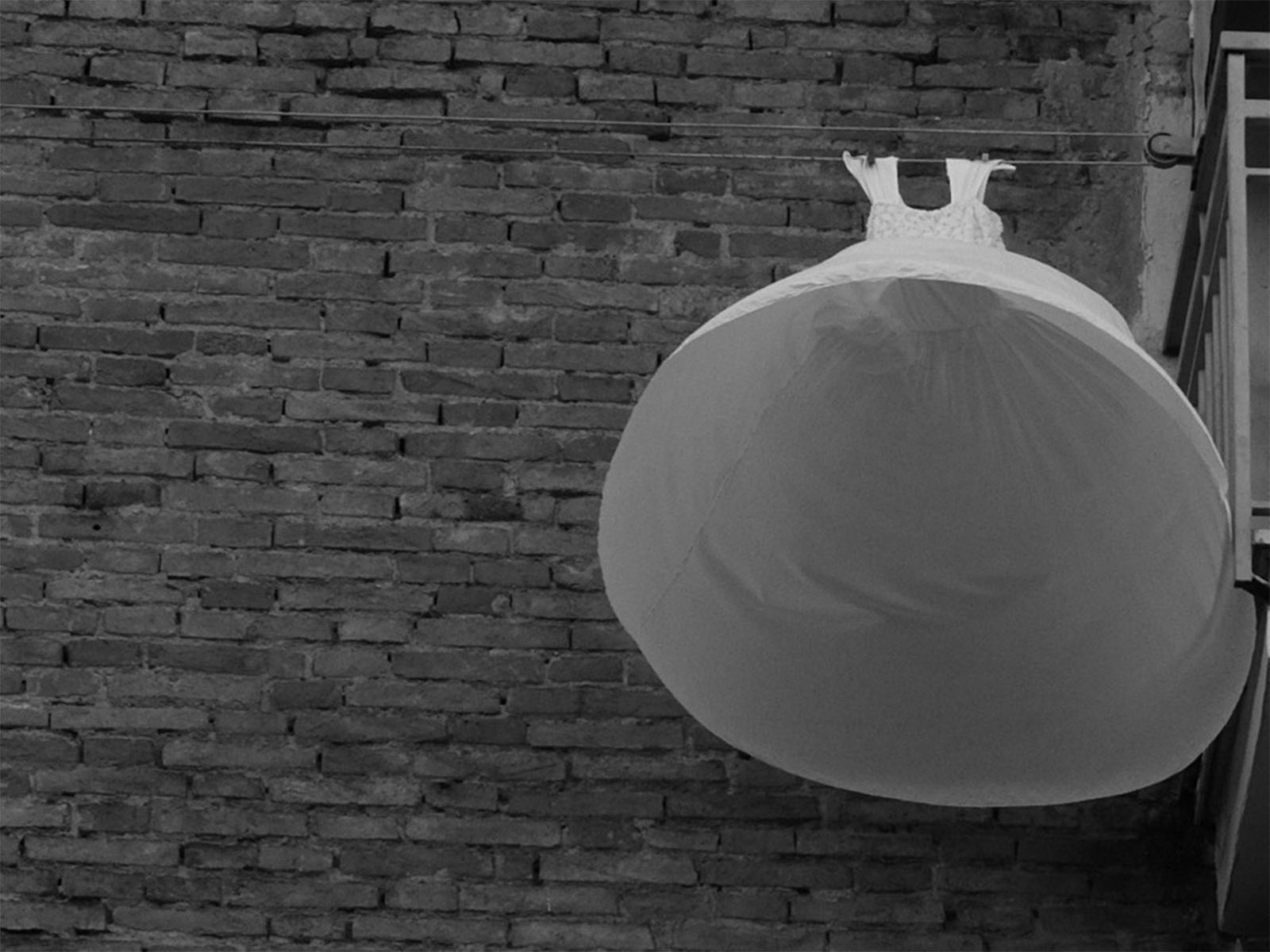 A white dress hangs on a clothesline next to a brick wall. The angle of the photo captures the dress from below, creating a balloon-like appearance. The scene is in black and white, emphasizing the contrast between the dress and the textured brick background.