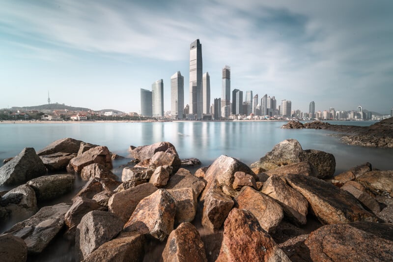 Cityscape viewed over a rocky foreground with a calm river reflecting skyscrapers under a clear sky.