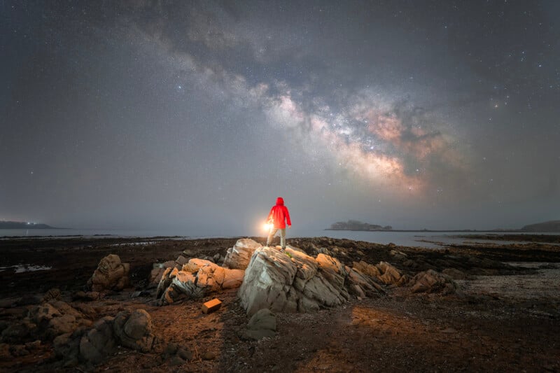 A person in a red jacket stands on rocky terrain under a starry sky, holding a bright light that illuminates the surrounding area and the milky way galaxy above.